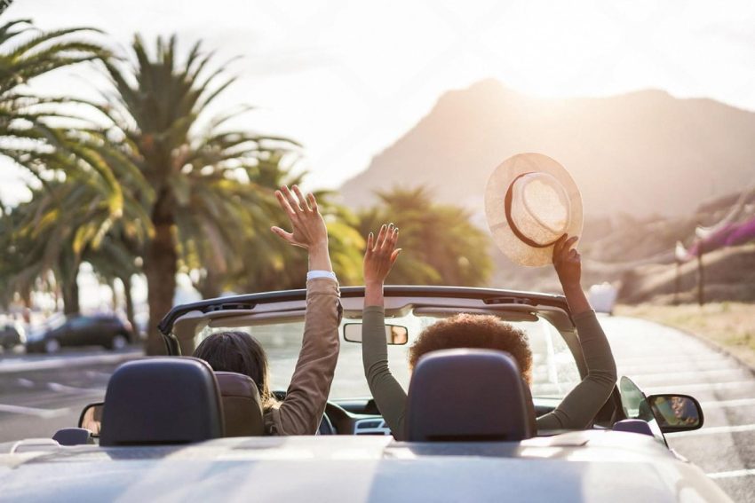 Two travellers in a convertible on a palm-lined Australian coastal road at sunset